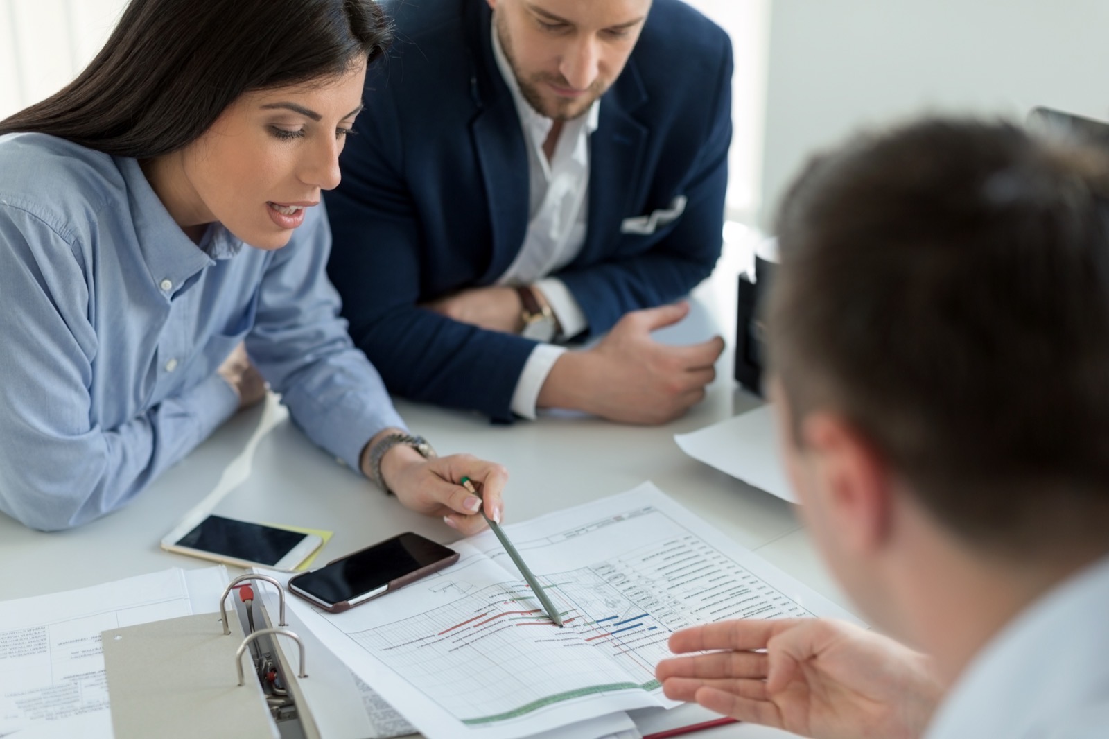 Three people engaged in a discussion over documents and charts at a table. One points with a pencil. Phones and binders are present.